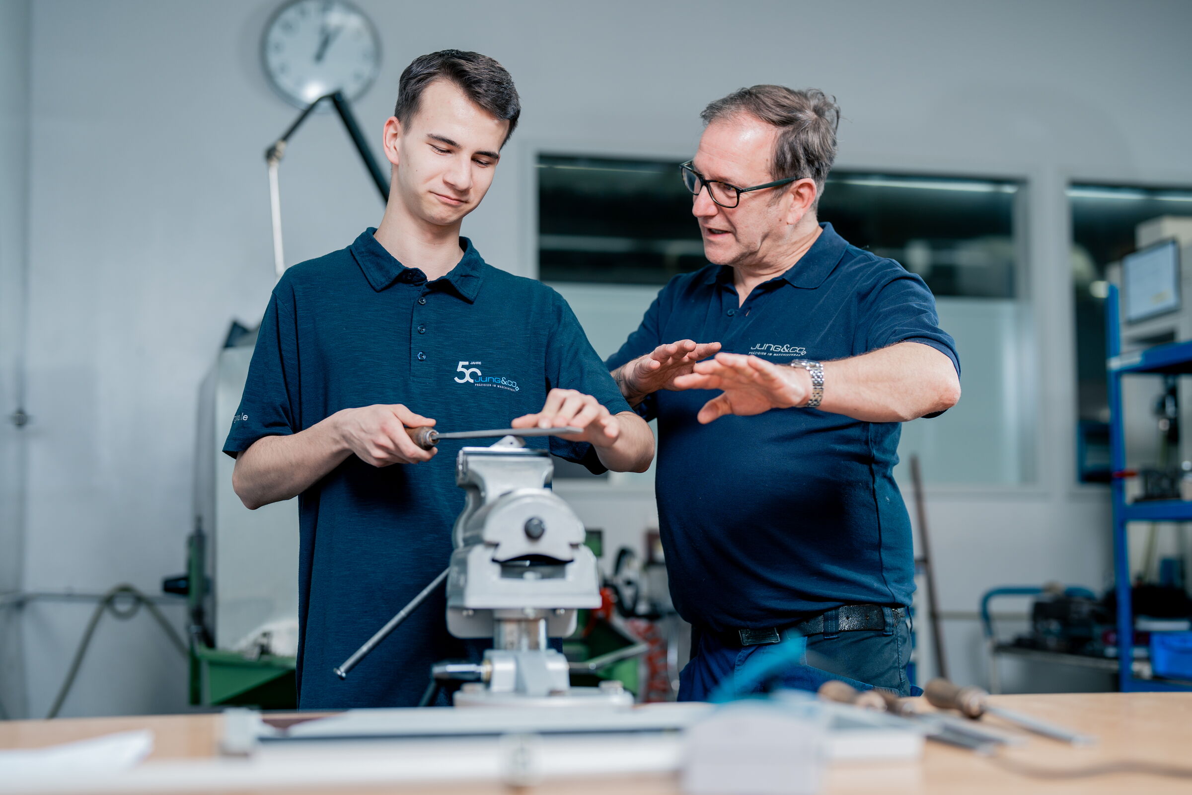 Zwei Männer bearbeiten ein Metallteil mit einer Feile. Sie befinden sich in einer Werkstatt