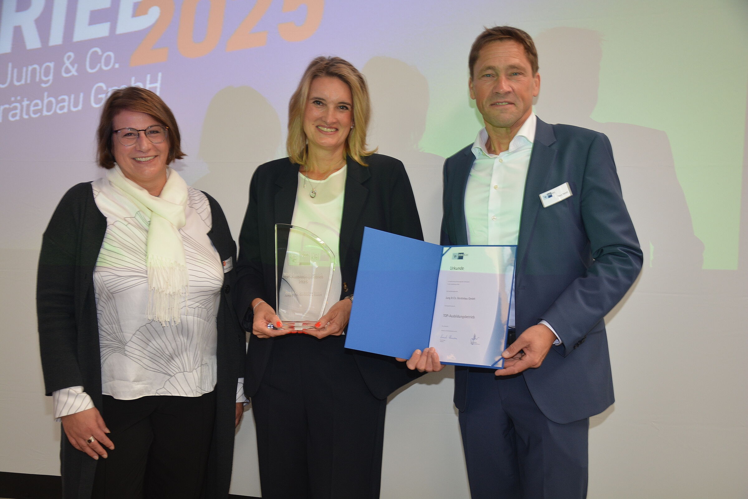 Zwei Frauen und ein Mann posieren vor einer Leinwand mit einem Pokal und einer Urkunde der IHK.