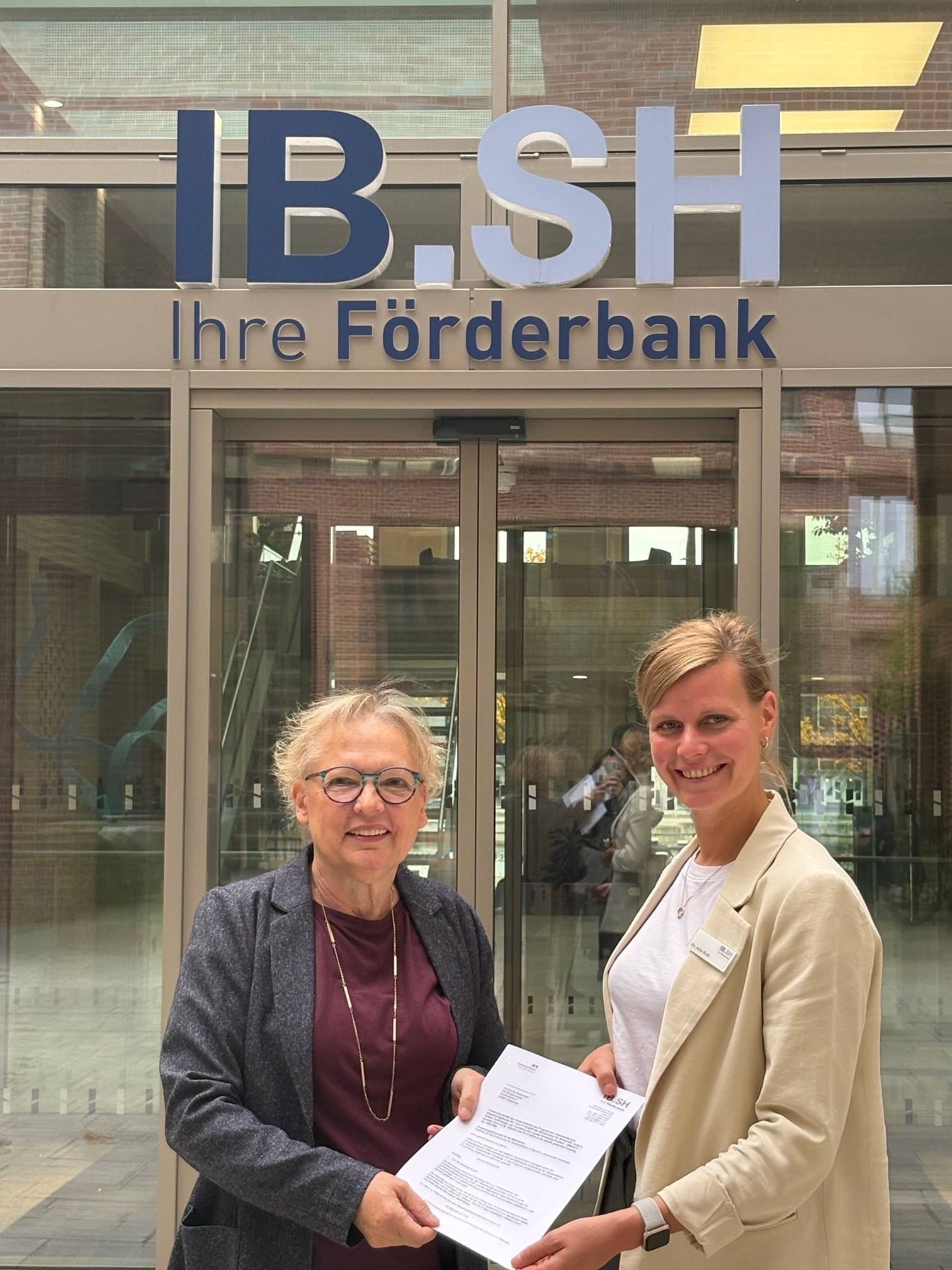 Zwei Frauen stehen vor dem IBSH Gebäude, sie präsentieren einen Zuwendungsbescheid.